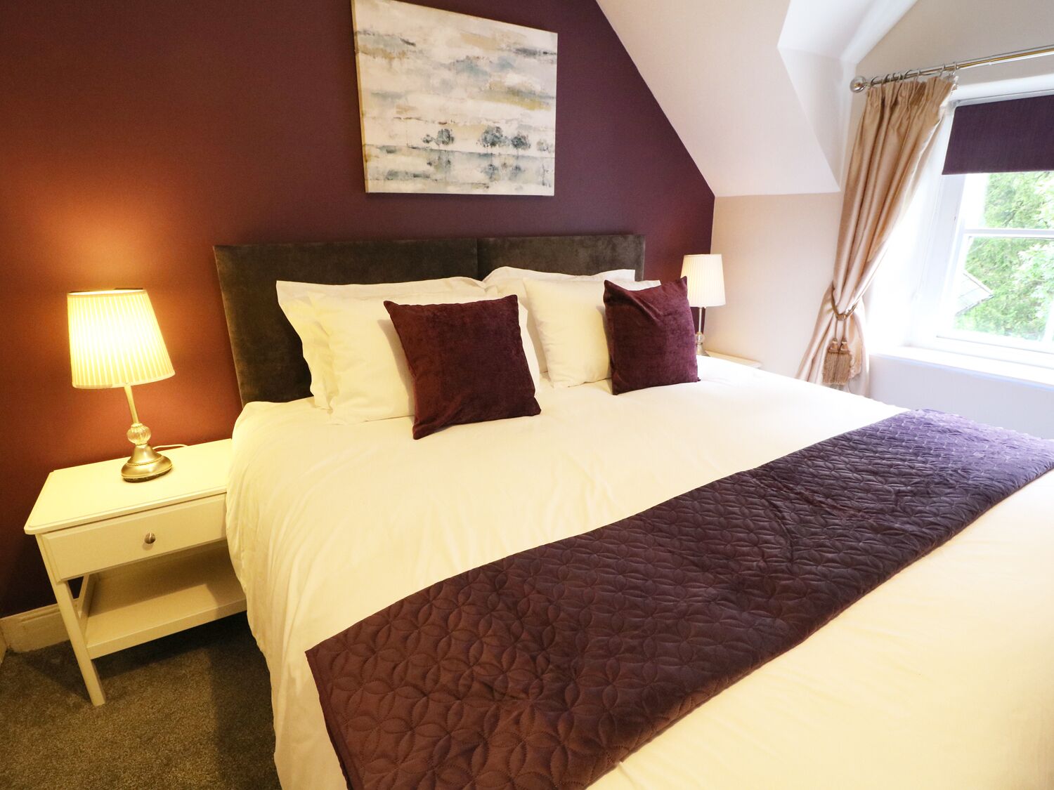A neatly made bed with a white and purple quilt, a dark headboard, and a bedside table with a lamp.