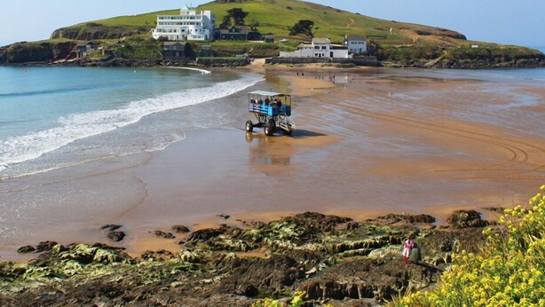 A coastal area with a beach, a hill with buildings, and a vehicle on the sand. - Homefield House (Kingsbridge)