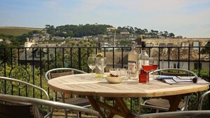 A round wooden table with metal chairs on a balcony, set with wine glasses, bottles, and a bowl of fruit.