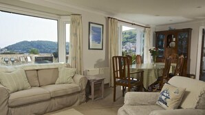 A living room with a sofa, armchair, and dining table. There is a view of a hill and a building through the windows.