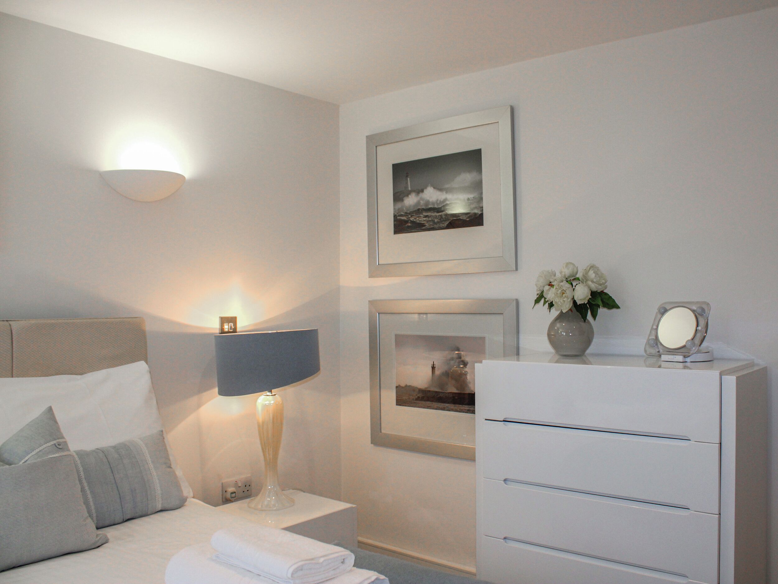 A neatly arranged bedroom with a bed, bedside lamp, framed pictures, and a dresser.