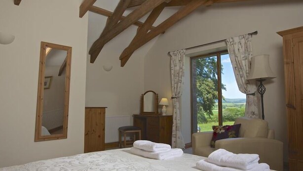 Ein Schlafzimmer mit einem großen Bett, Holzbalken an der Decke, einem Fenster mit Blick ins Grüne und einem Spiegel an der Wand.