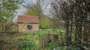 Une maison de campagne en briques, avec un toit recouvert de tuiles rouges, une porte en bois et un jardin où poussent différentes plantes et fleurs.