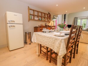 Un comedor con una mesa blanca dispuesta para cuatro personas, sillas de madera, un refrigerador blanco y una estantería de madera con diversos objetos.