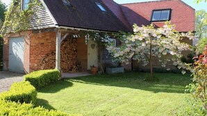 Une maison de campagne au toit en pente, avec une véranda en bois et un jardin agrémenté d’un arbre en fleurs.