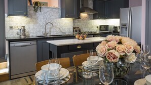 A modern kitchen with dark cabinets, a marble backsplash, and a dining area set for a meal. - 16 Courtenay Street (Salcombe)
