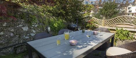 Un patio al aire libre con una mesa lista para comer, rodeado de sillas de mimbre y un muro de piedra.