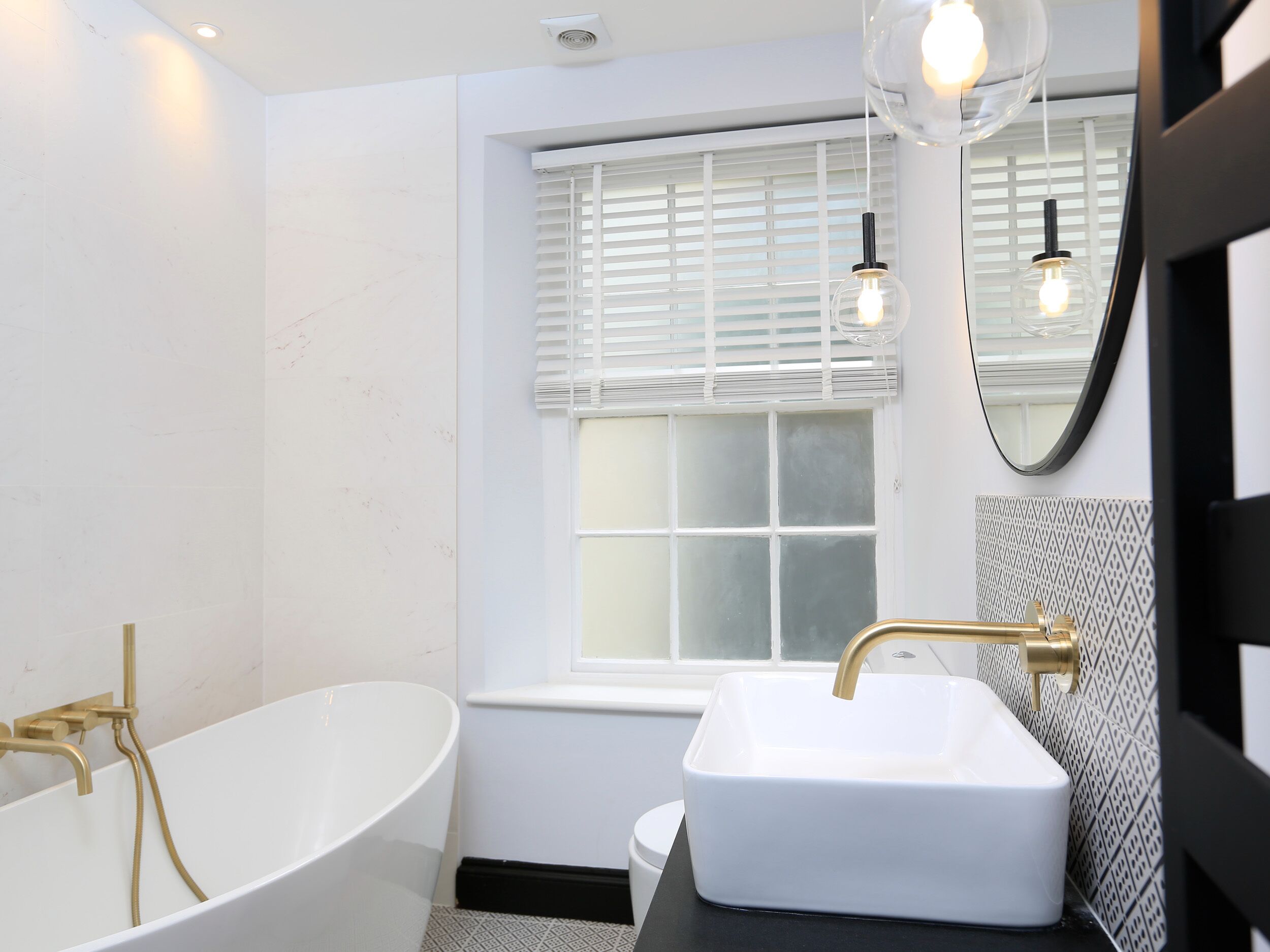 A modern bathroom with a freestanding bathtub, a white sink with a gold faucet, and a window with blinds.