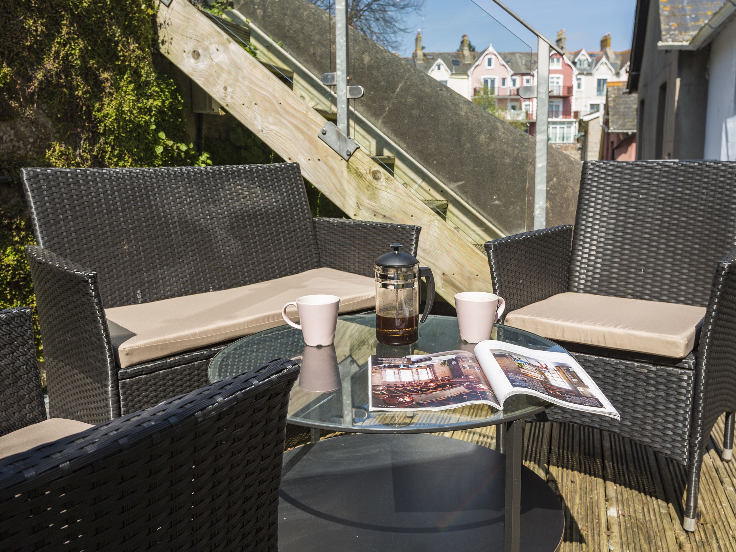 A wicker outdoor seating area with a glass table, two chairs, a coffee pot, and a magazine.