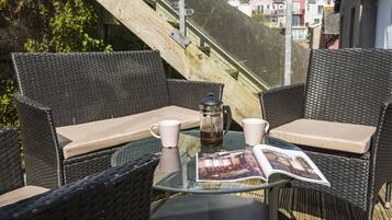 A wicker outdoor seating area with a glass table, two chairs, a coffee pot, and a magazine.