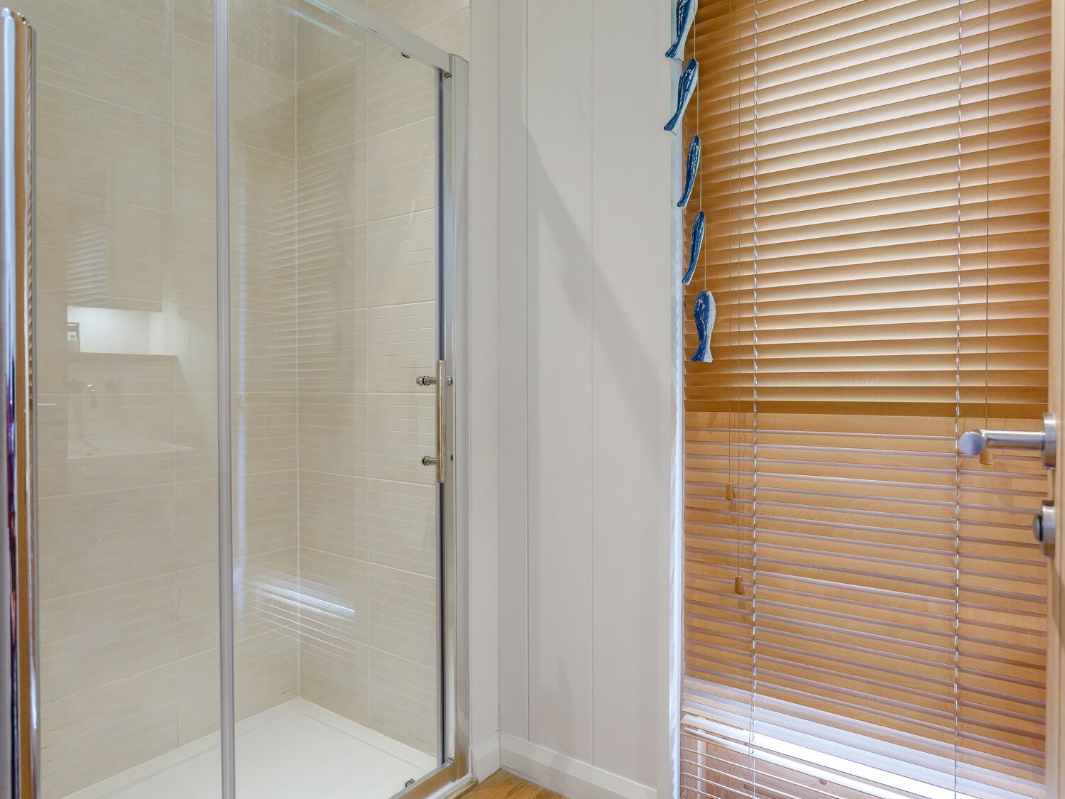 Un espace douche entièrement vitré, avec un mur recouvert de carreaux blancs et une porte en bois équipée de stores.