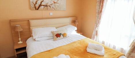 A neatly made bed with white linens and a yellow blanket, a painting of birds on a tree, and a window with sheer curtains.