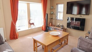 Een woonkamer met een houten tafel, oranje gordijnen, een wandtelevisie en een boekenkast.