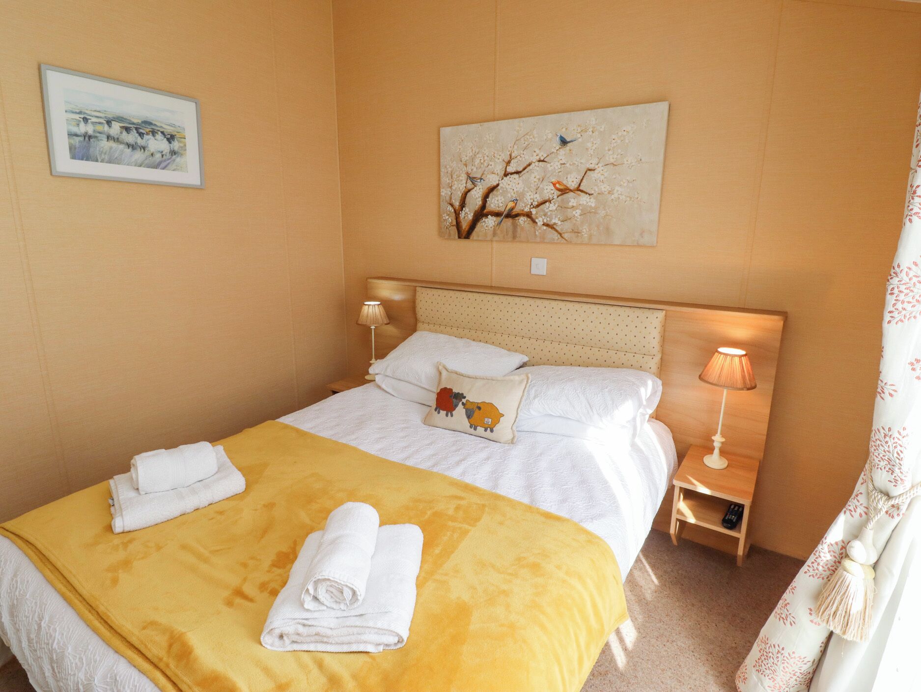 A neatly made bed with white linens, a yellow throw blanket, and two rolled towels. There is a small bedside table with a lamp and a remote control.