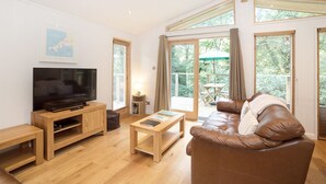 A modern living room with a flat-screen TV, wooden furniture, a leather sofa, and large windows offering a view of the outdoors. - 7 Streamside (Looe)