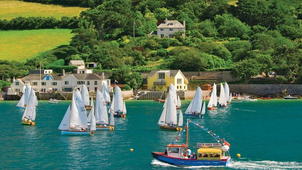 Un paysage côtier avec des voiliers sur l’eau, un ferry et des maisons sur le versant de la colline.