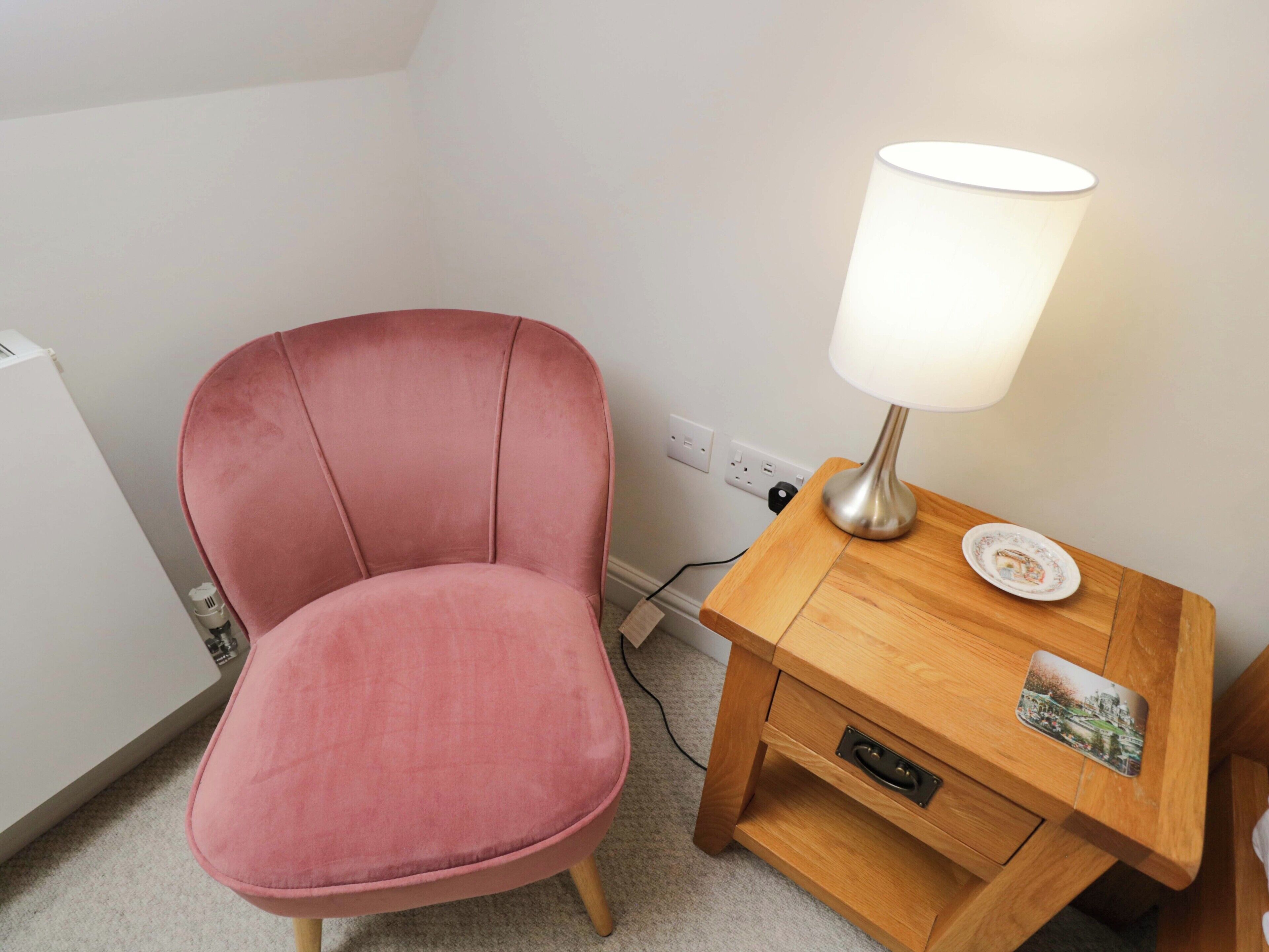 Une chaise rose est placée à côté d’une table de chevet en bois, sur laquelle se trouvent une lampe et une petite assiette.