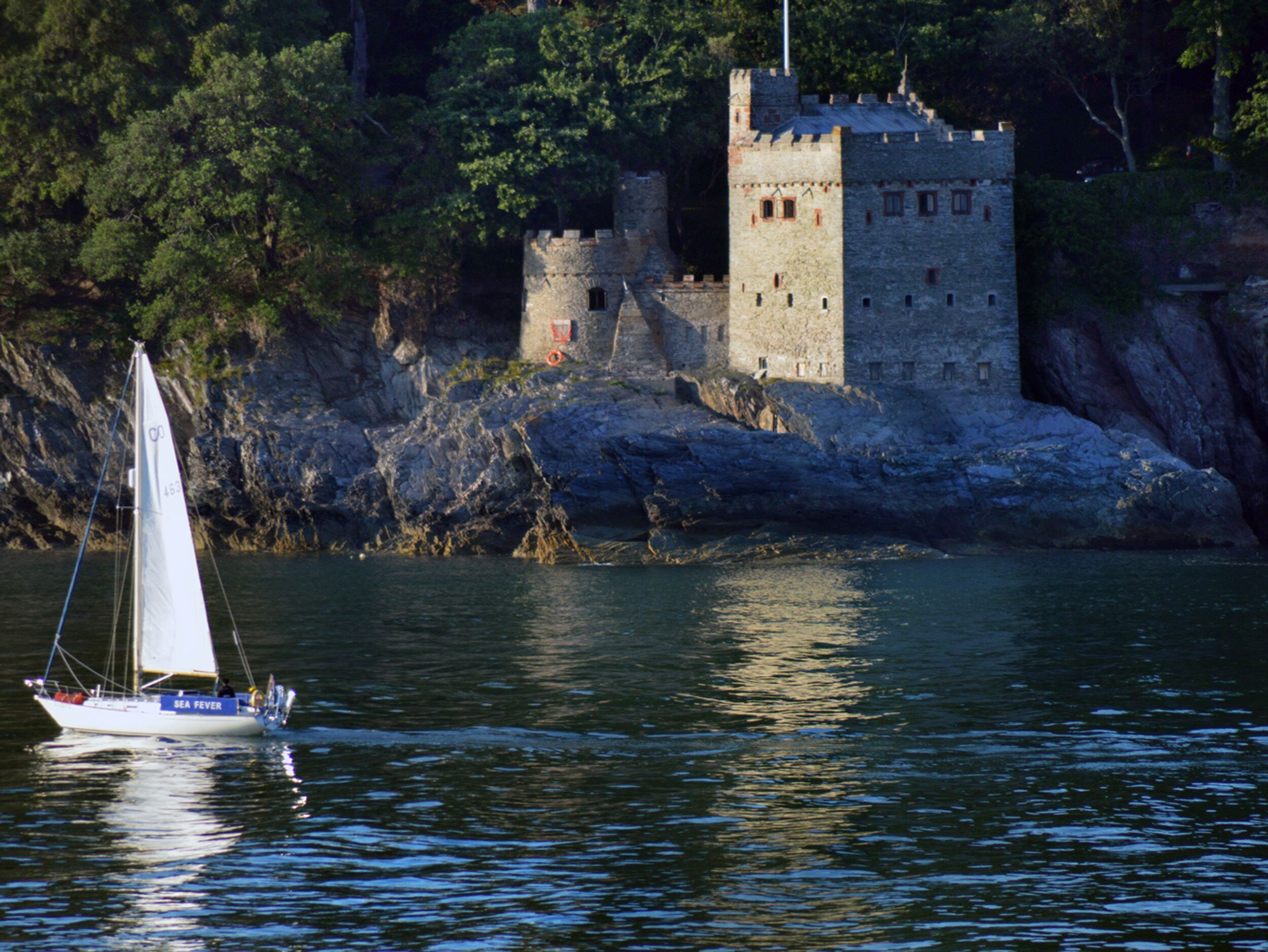 Un voilier sur une étendue d’eau calme, près d’un château de pierre historique entouré d’arbres.