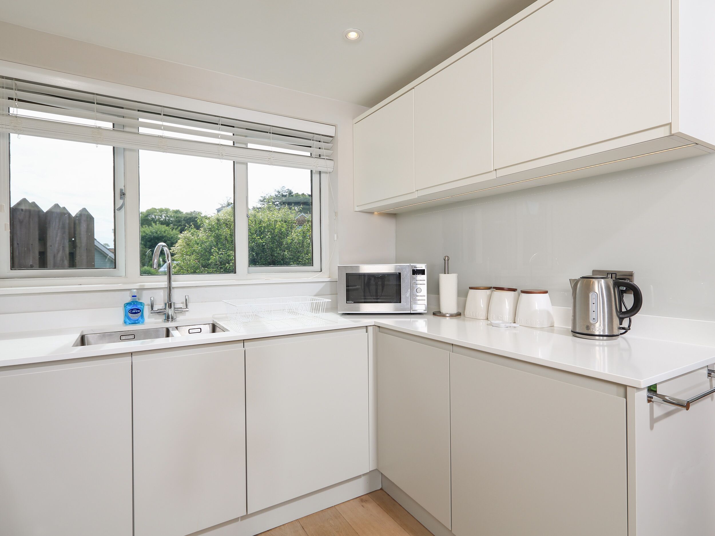 Una cocina moderna con armarios blancos, microondas integrado, hervidor y ventana con persianas.