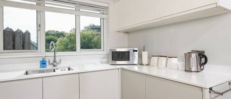 Una cocina moderna con armarios blancos, microondas integrado, hervidor y ventana con persianas.