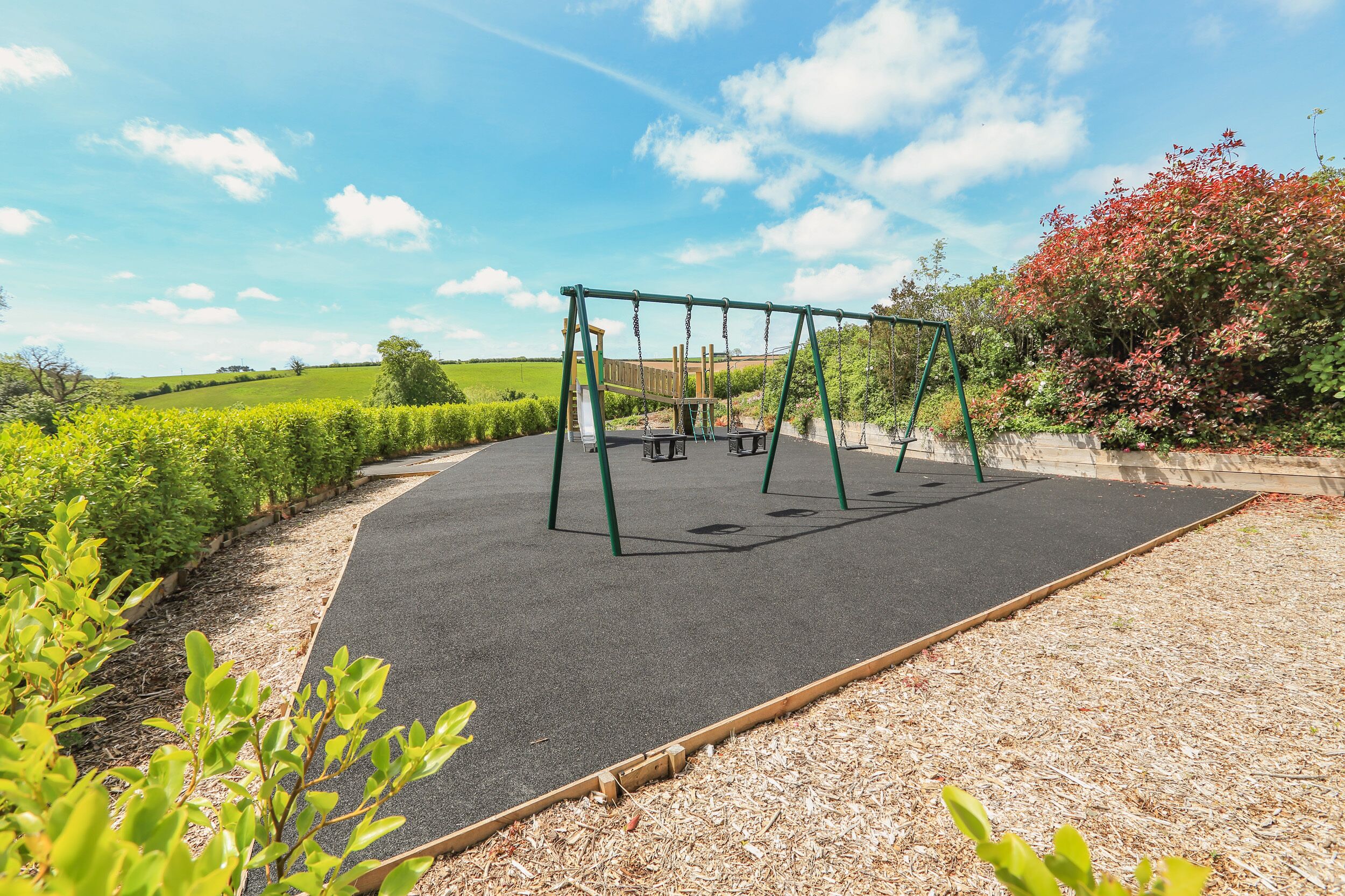 A playground with swings and a slide, surrounded by greenery and a clear sky.