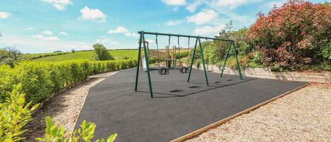 A playground with swings and a slide, surrounded by greenery and a clear sky.