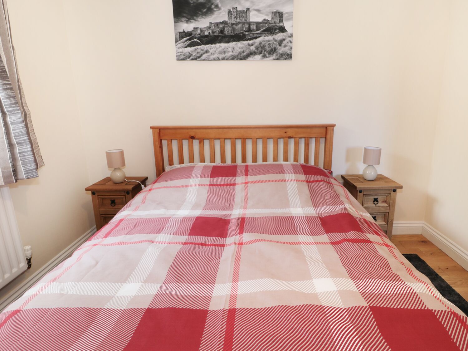Ein Schlafzimmer mit einem hölzernen Bett, einer karierten Bettdecke, zwei Nachttischen mit Lampen und einem Wandbild eines Schlosses.