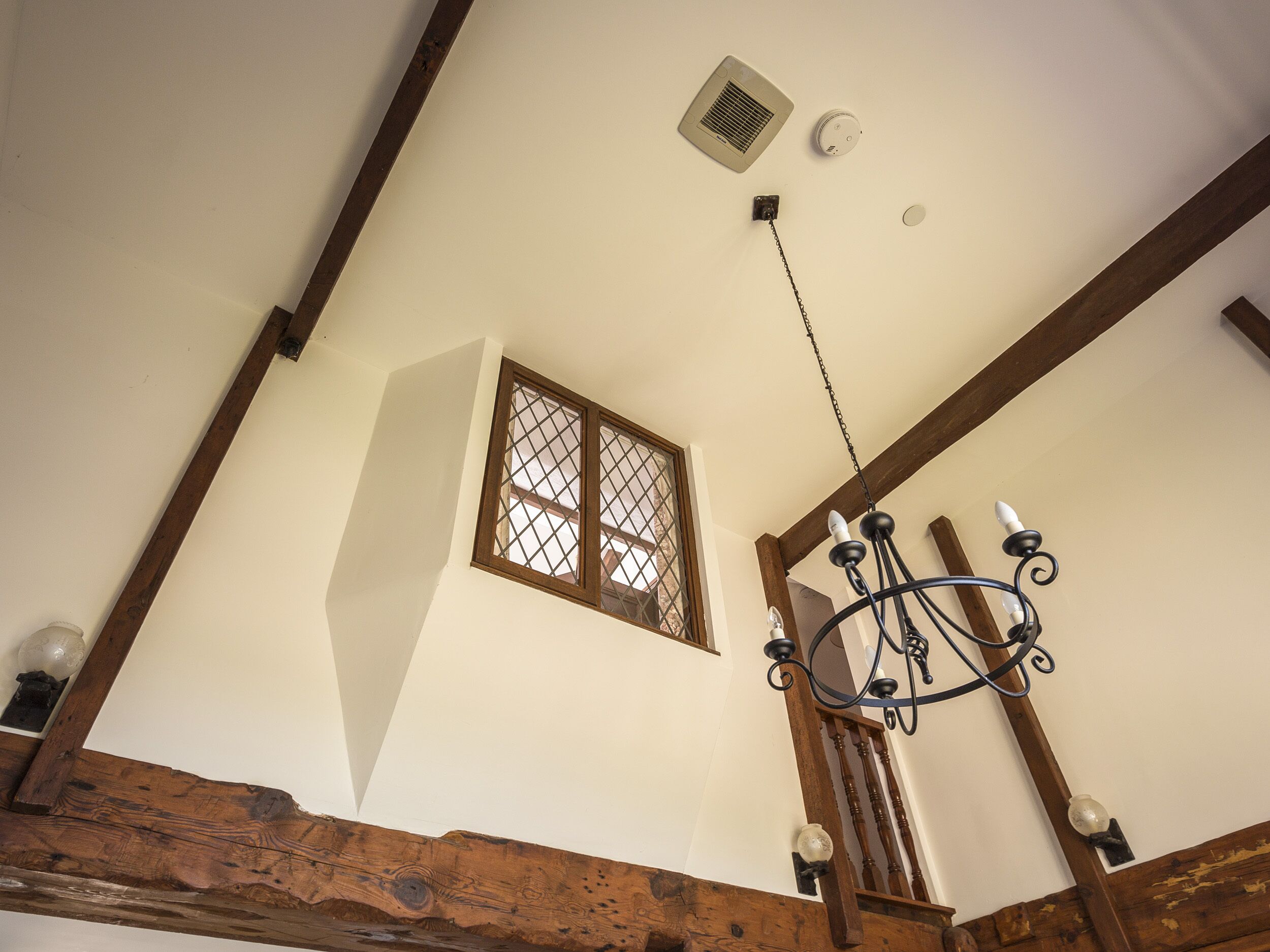 A high ceiling with a wooden beam structure, a window with a grid pattern, and a chandelier.