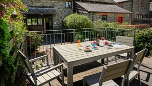 Een houten tafel met stoelen en een metalen hek in een tuin, met stenen gebouwen op de achtergrond.