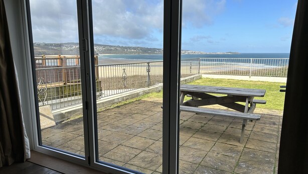 Vue depuis une chambre dotée d’une grande porte-fenêtre coulissante donnant sur un patio extérieur avec une table de pique-nique et une rambarde en métal, d’où l’on aperçoit une plage.