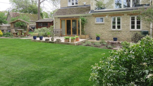 A two-story stone house with a covered patio, a lawn, and various potted plants.