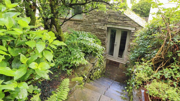 A stone building with a door, surrounded by lush greenery and a small stone staircase. - Holemire Barn (Cockermouth)
