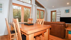 Una mesa de madera con sillas, un sofá y un televisor en una habitación con ventanas amplias y vistas a árboles.