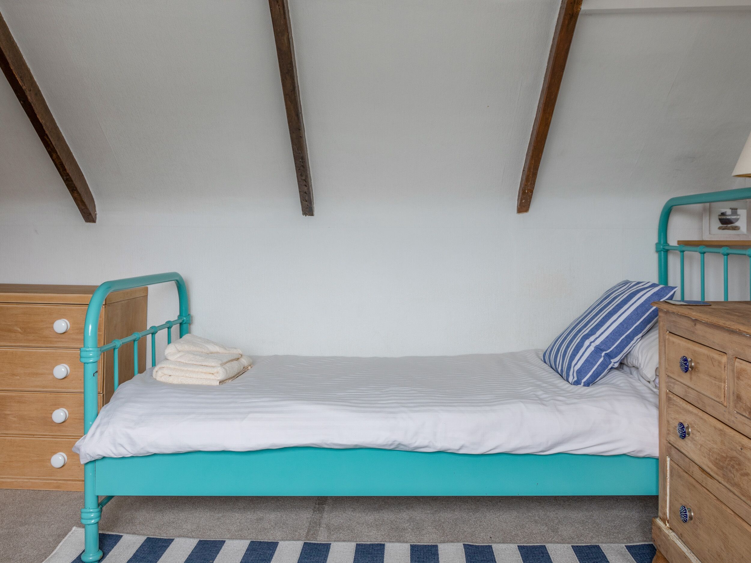 Una cama individual con estructura color turquesa, ropa de cama blanca y almohada a rayas.