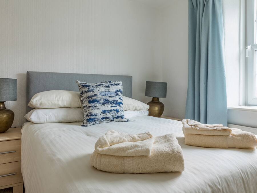 Un lit bien fait, avec des draps blancs, un oreiller à motifs bleus et blancs, et deux serviettes pliées posées sur le lit.