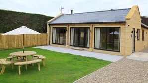 A single-story house with a sloped roof, large glass windows, and a patio area with a wooden table and chairs. - Park Bothy (Cheltenham)