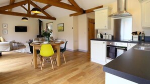 A spacious living area with a wooden ceiling, a dining table with chairs, a kitchen with white cabinets and a stainless steel range hood. - Park Bothy (Cheltenham)