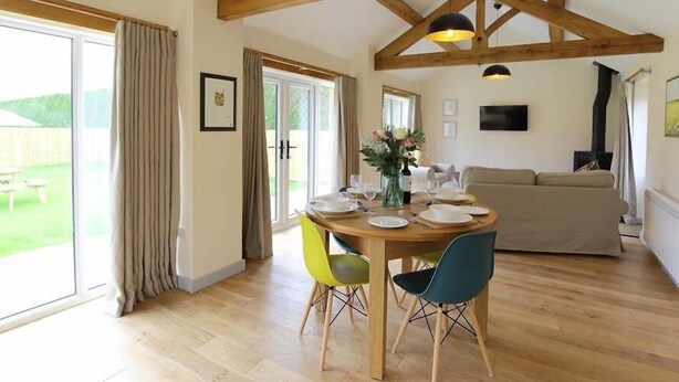 A dining area with a wooden table set for four, surrounded by chairs. There is a large window with curtains, a sofa in the background, and a fireplace. - Park Bothy (Cheltenham)