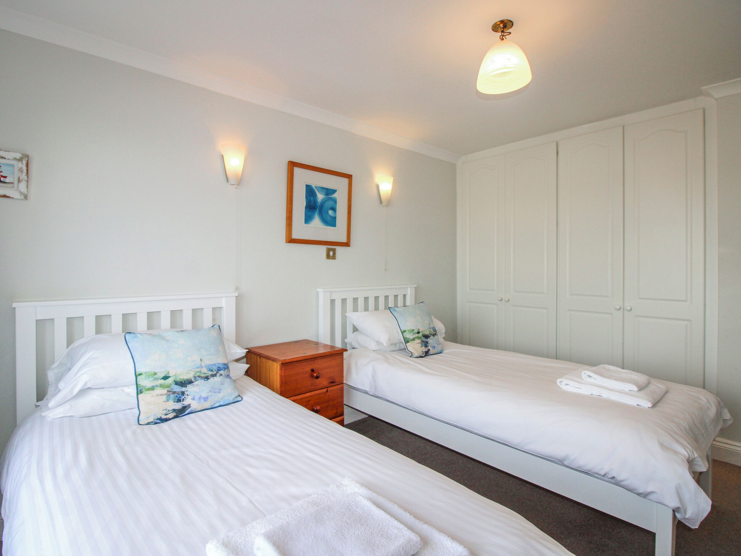 Une chambre avec deux lits simples, une table de chevet en bois, un luminaire fixé au mur, une porte et un tableau encadré accroché au mur.