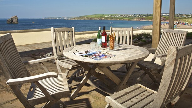 Une table en bois avec des chaises, une bouteille de vin et un verre d’eau, le tout sur une terrasse donnant sur une plage.