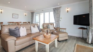 Un salon avec un canapé beige, une table basse en bois, un téléviseur et un coin repas comprenant des chaises et une table.