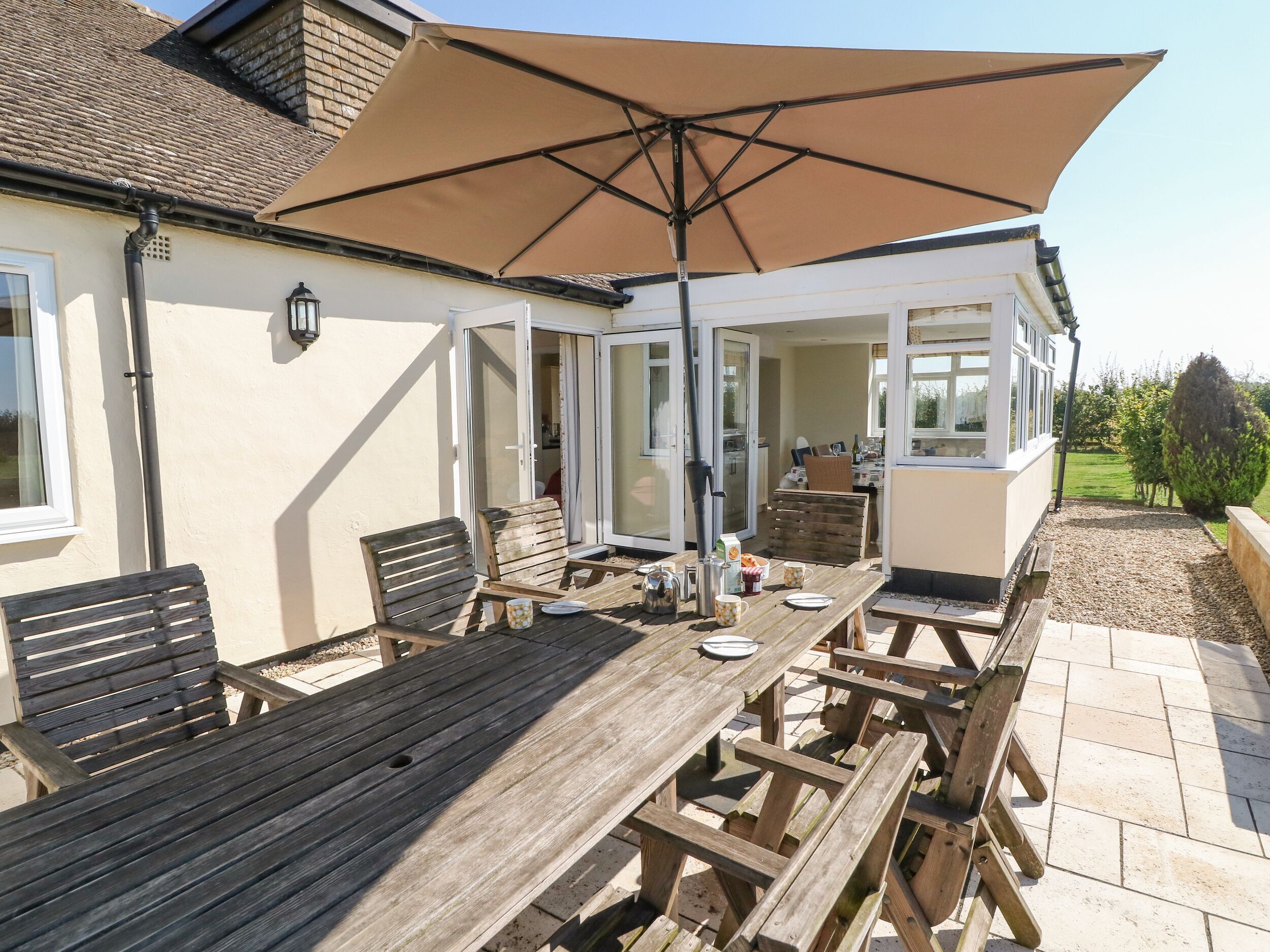 A patio area with a wooden table and benches, a large umbrella, and a view of the house's interior through glass doors.