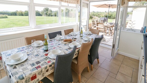 Un coin repas avec une table dressée pour quatre personnes, des chaises en osier, une nappe à motifs et une vue sur un jardin grâce à de grandes fenêtres.