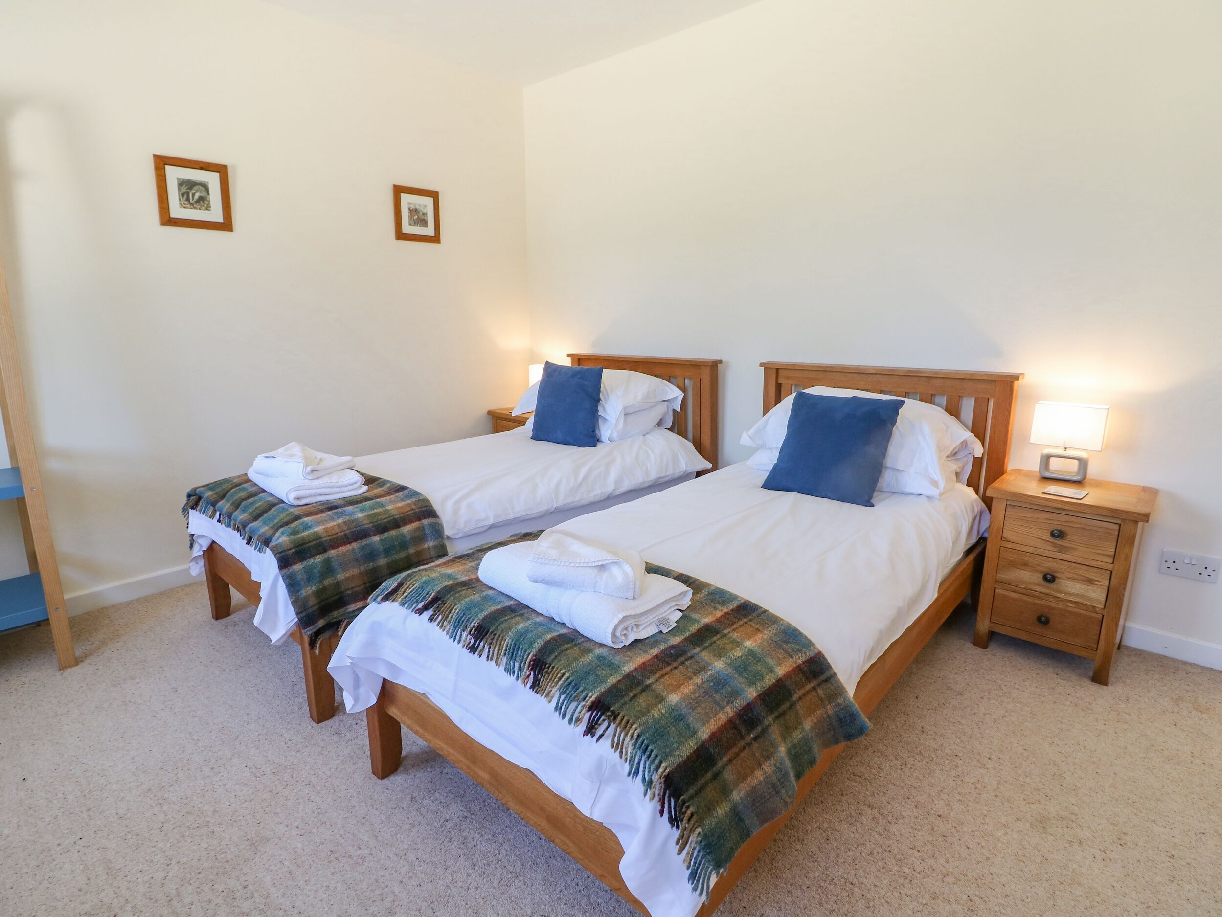 A hotel room with two single beds, each with a wooden headboard, white bedding, and a plaid blanket. There are two bedside tables with lamps, and the room has a carpeted floor.