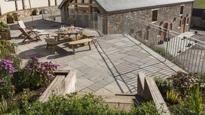 A stone building with a sloped roof, a patio with wooden furniture, and a garden area with flowers. - Butterwell Barn (Dartmouth)