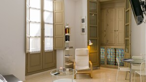 A room with a sofa, a glass table with candles, a white chair, a small table with a lamp, and a cabinet with books. - Very bright apartment in the historic center - MALAMA B2  (Málaga)
