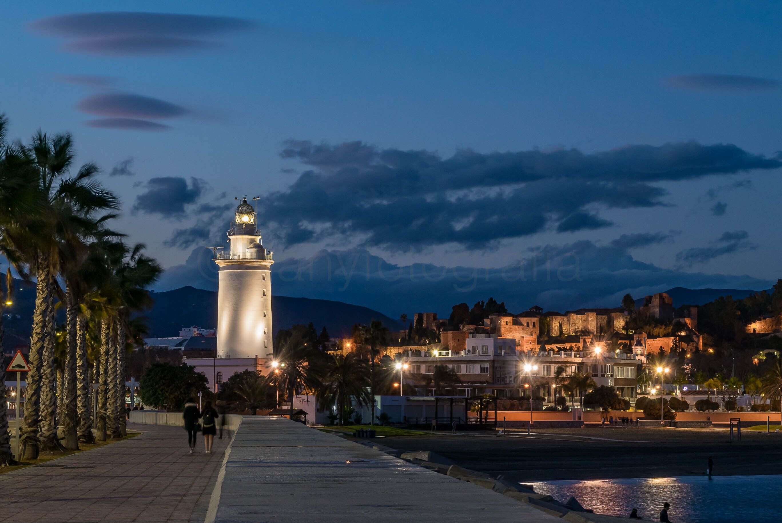 Une ville côtière au crépuscule, avec un phare imposant, des palmiers et des bâtiments illuminés.