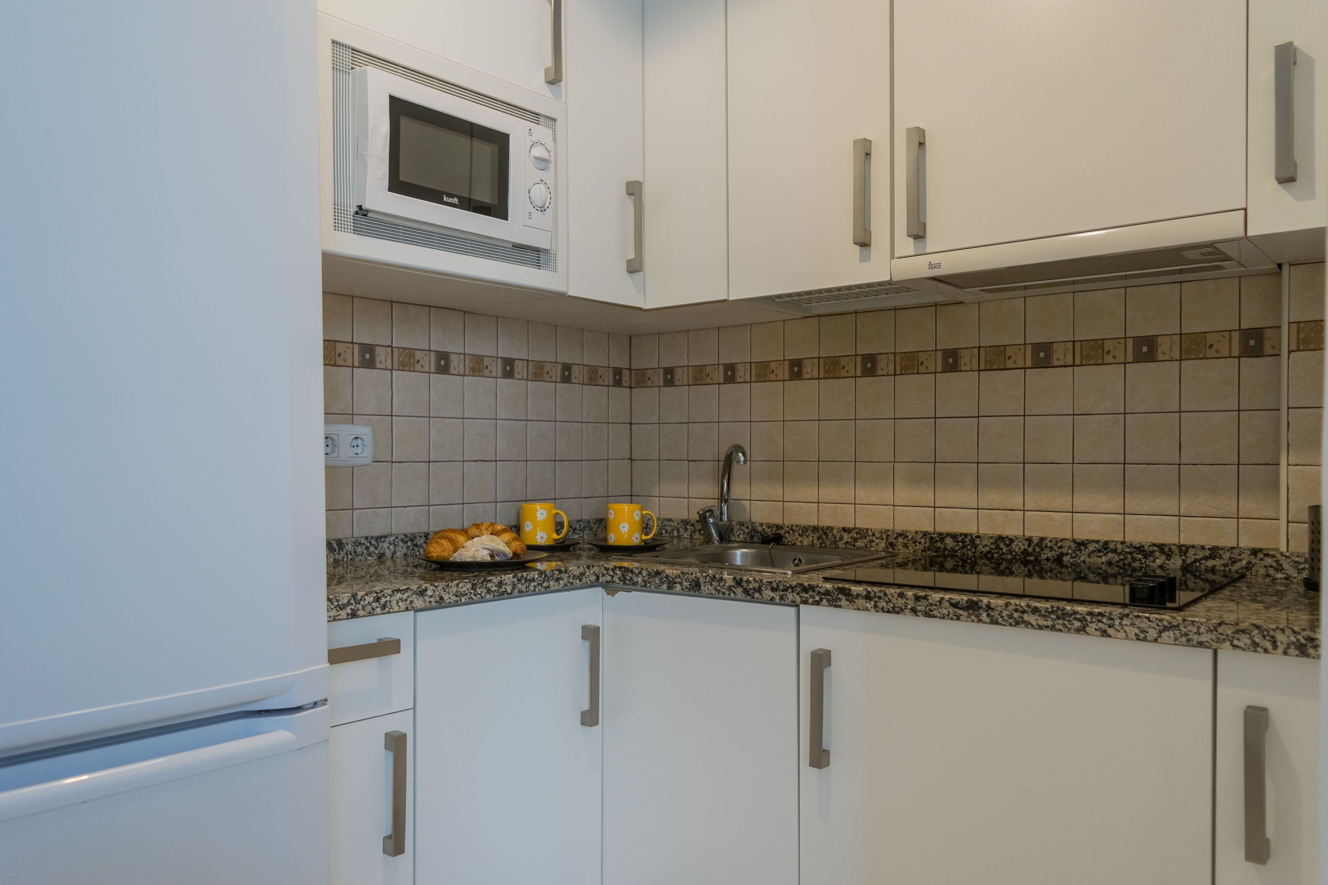 Una cocina con armarios blancos, microondas, lavamanos y encimera de granito.