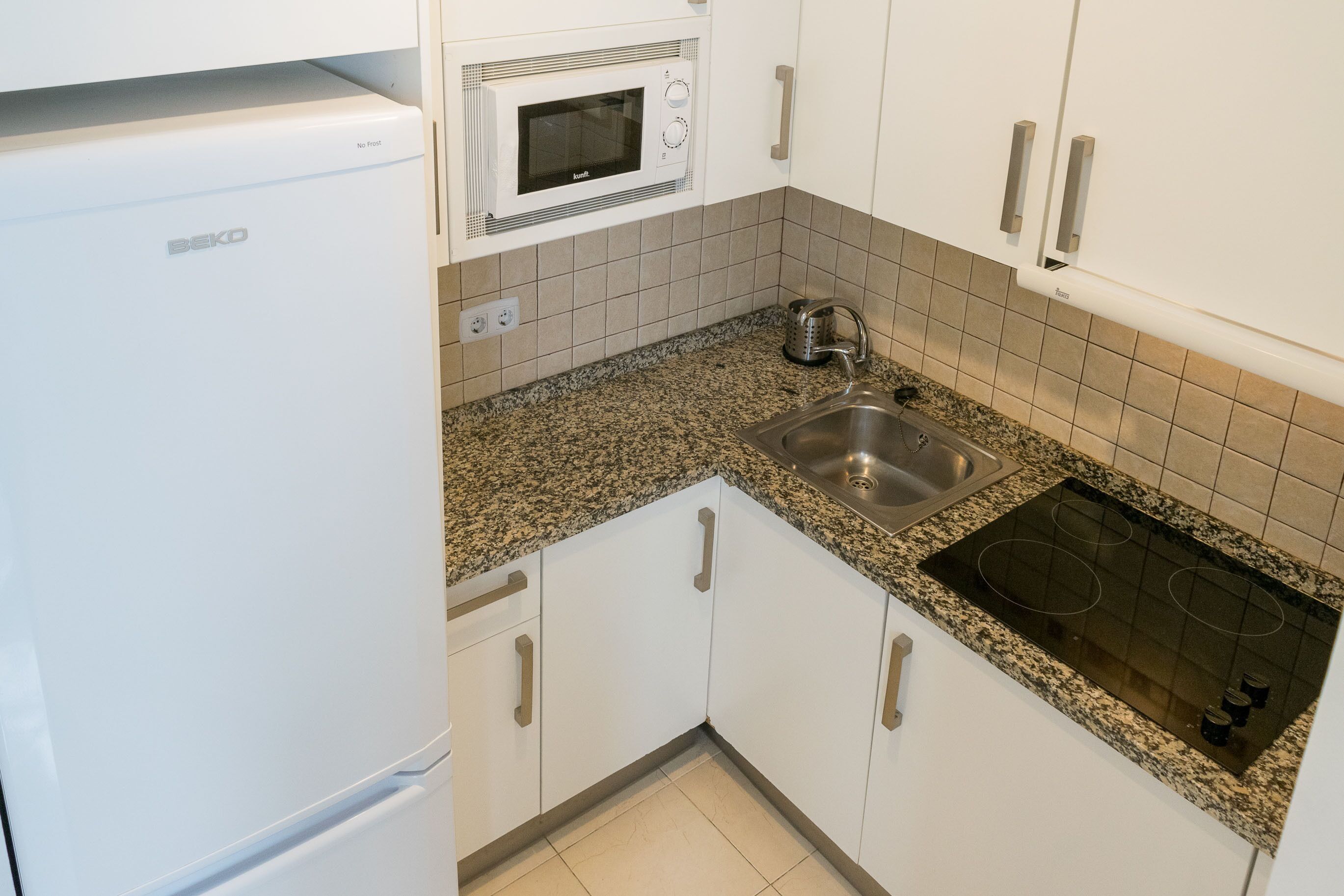 A compact kitchen with white cabinets, a built-in microwave, a stainless steel sink, and a stove.