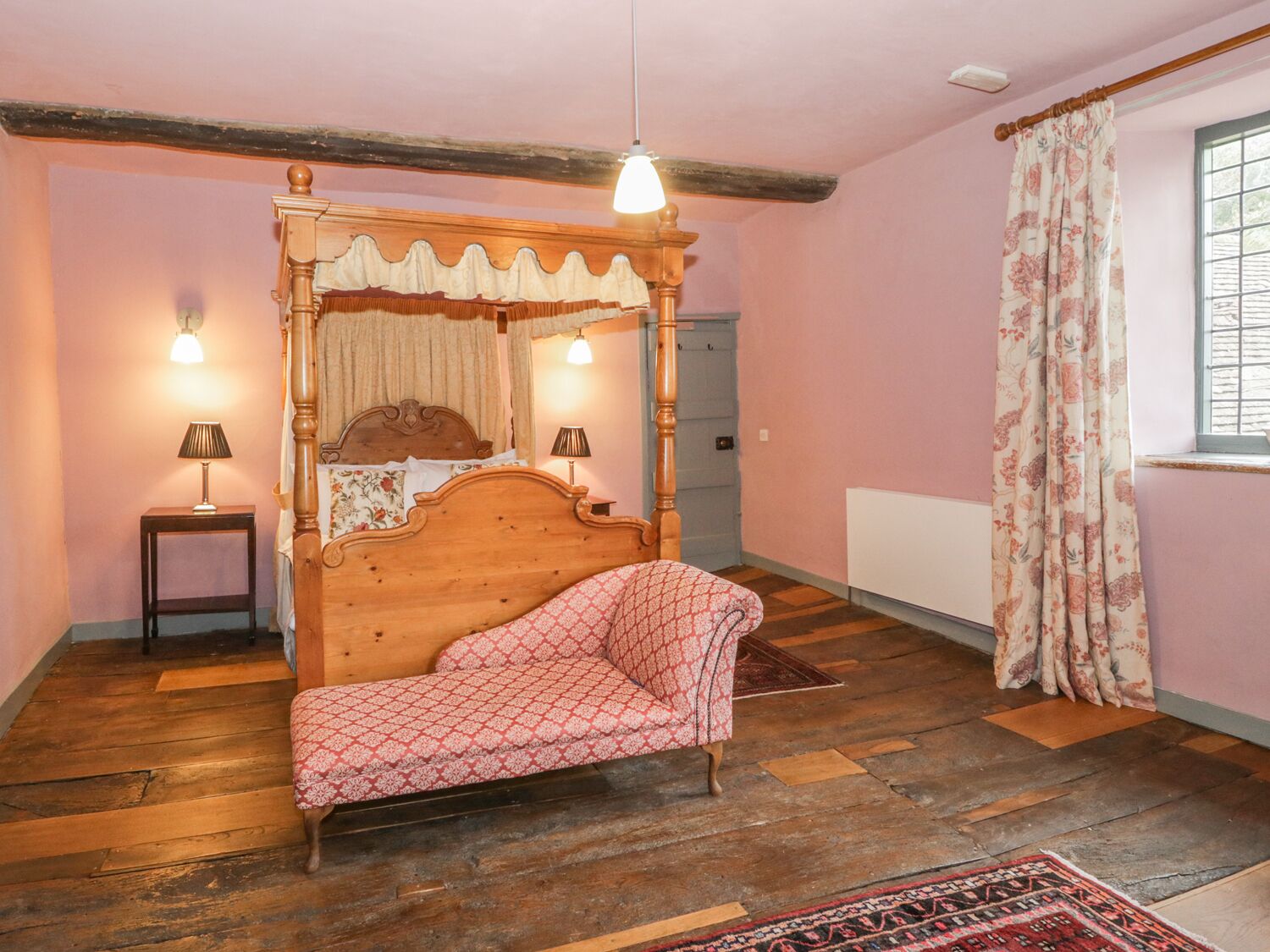 A bedroom with a four-poster bed, two bedside lamps, a wooden floor, a window with floral curtains, and a radiator.
